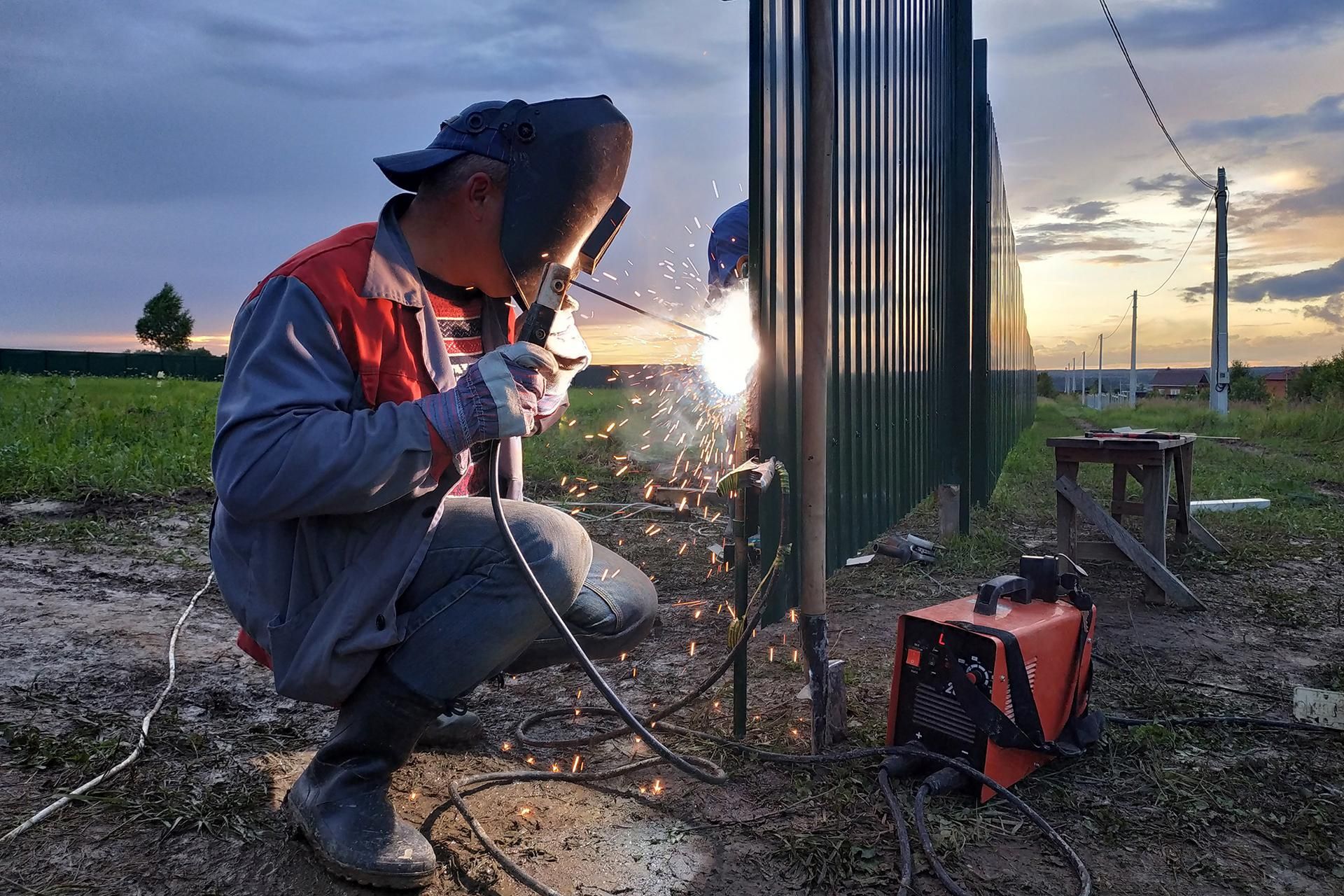 A man wearing a welding mask is welding a metal fence.