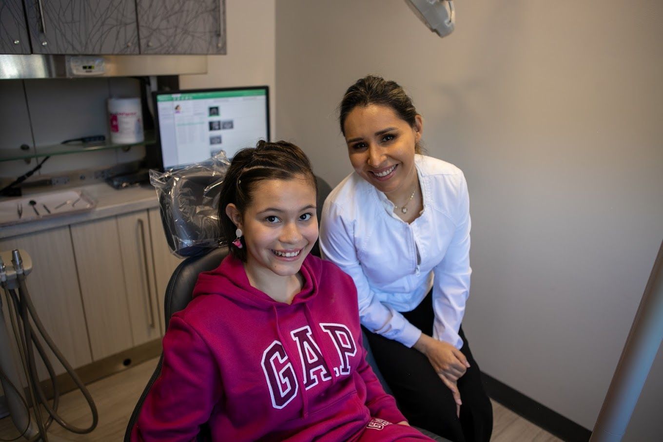 A girl in a gap sweatshirt is sitting in a dental chair next to a dentist.