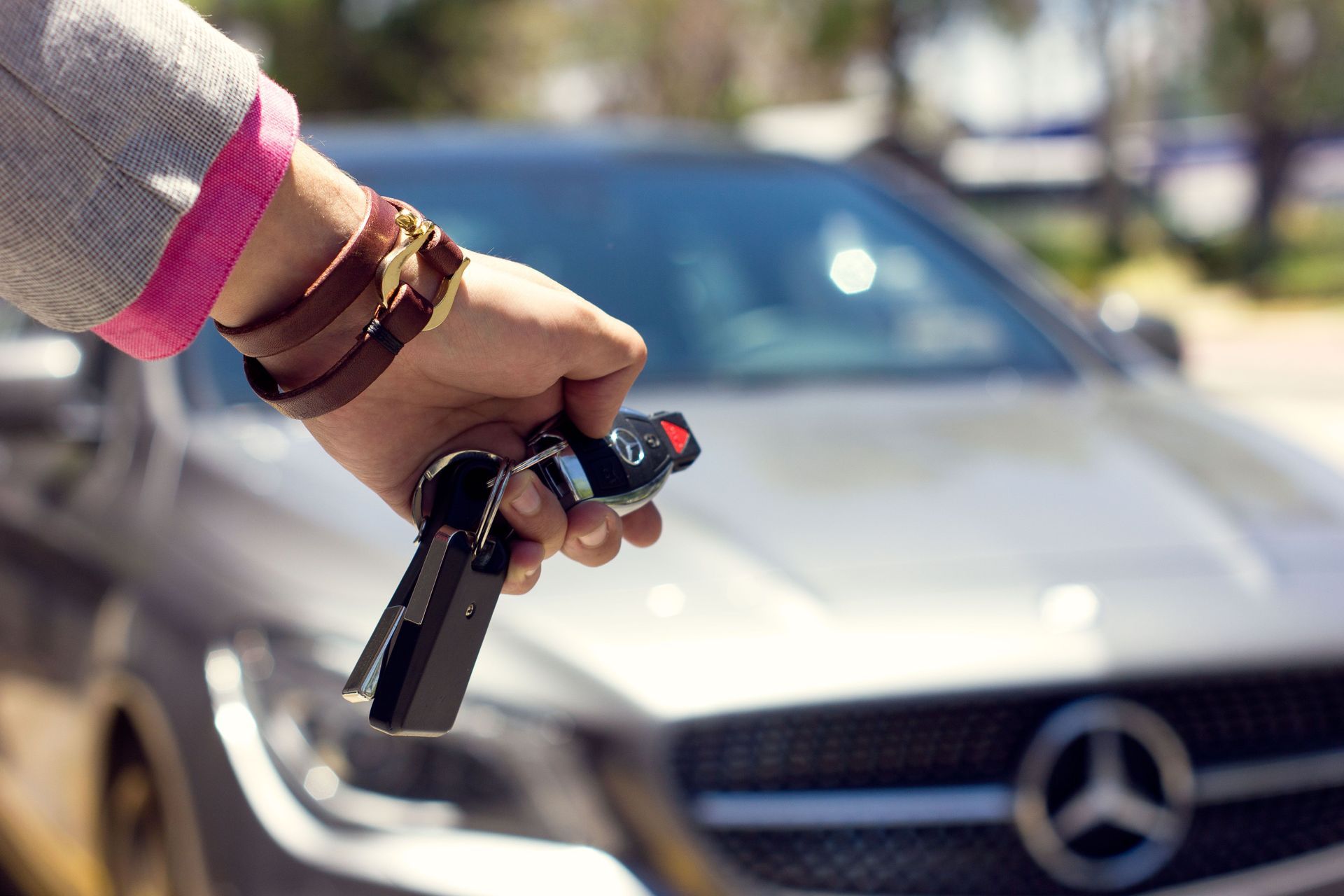 Hand holding car key fob in front of a silver Mercedes car outdoors