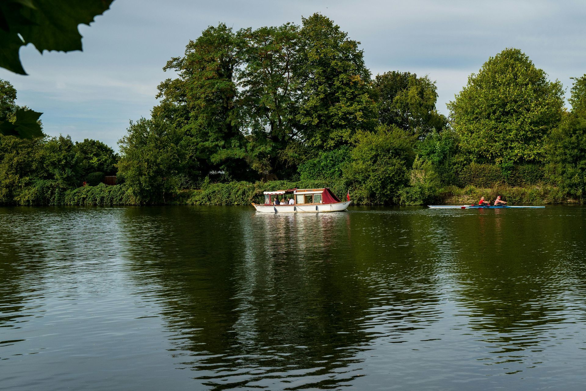 Boat on a calm river with trees lining the banks, two people rowing in the distance.