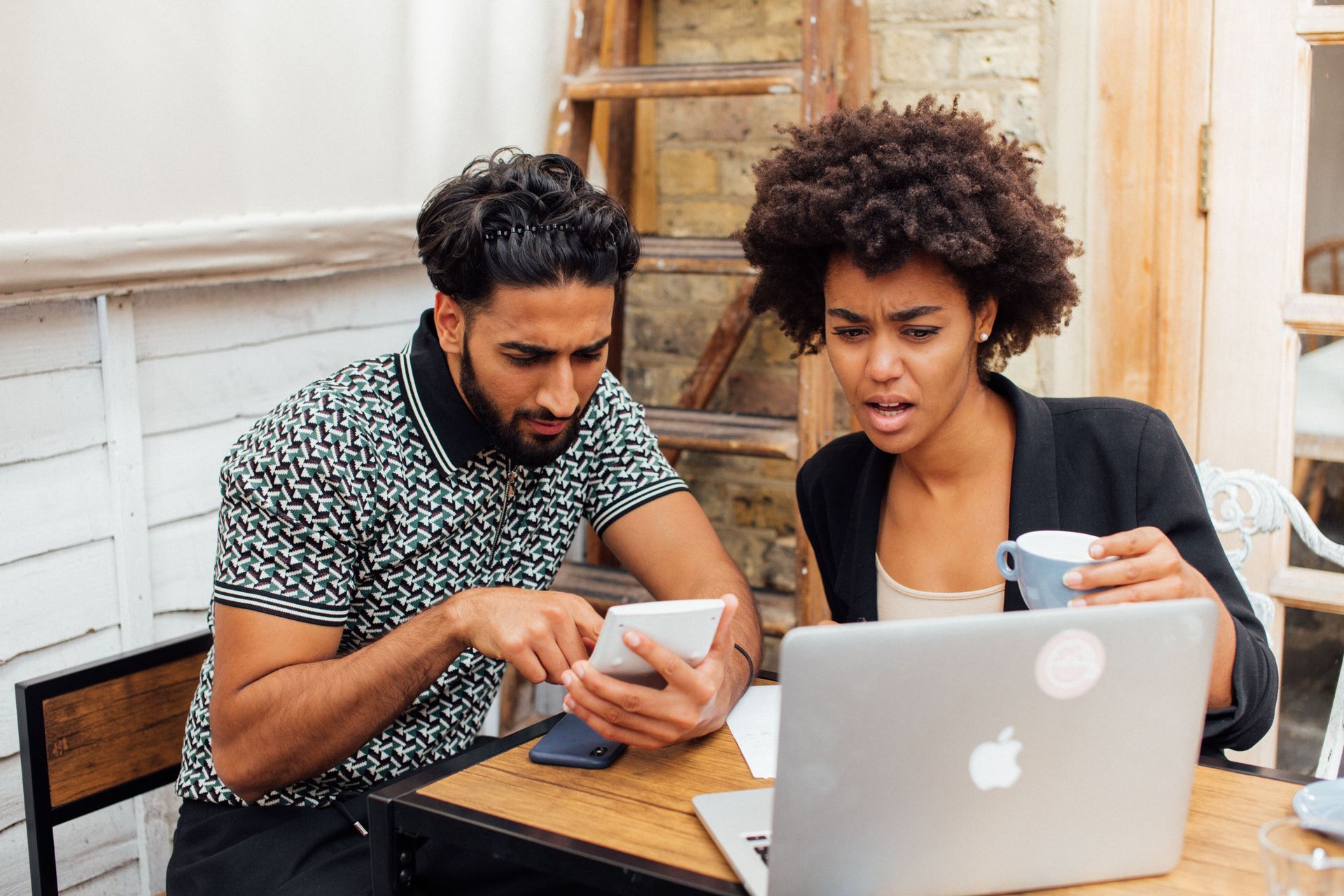 Two people working on a laptop and smartphone at a table indoors, looking focused.