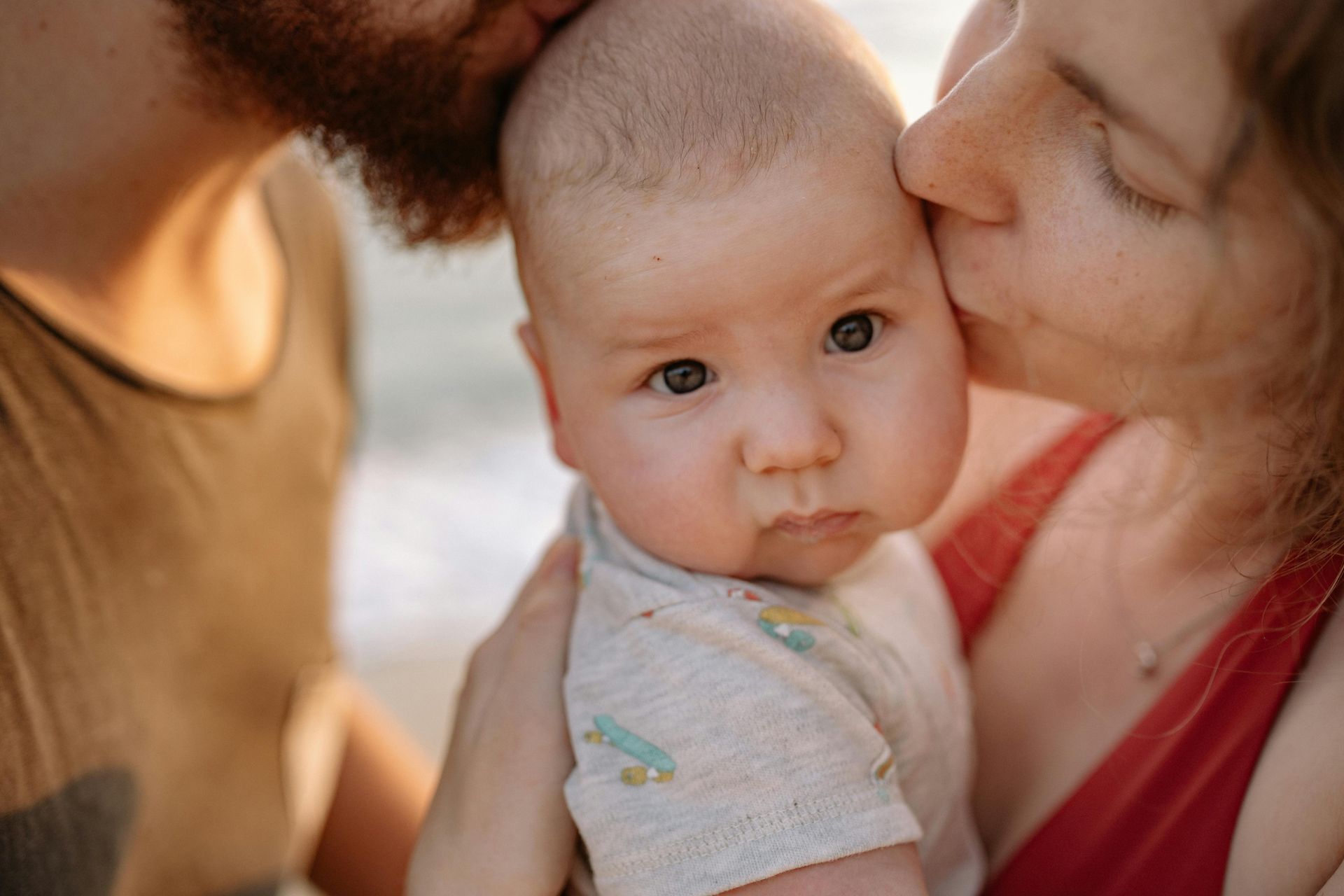 Parents kissing a baby's head; outdoor setting with soft light. Baby looks at the camera.