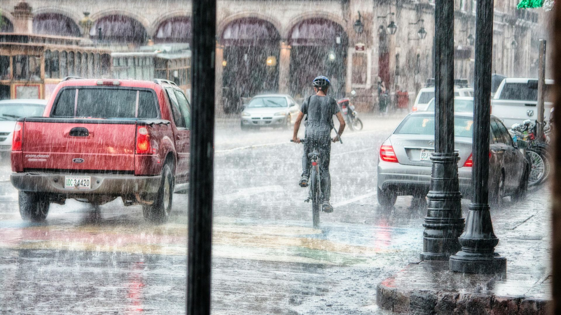 Rainy street scene with a cyclist, red pickup truck, and cars driving past buildings with arches and Mexican flags.