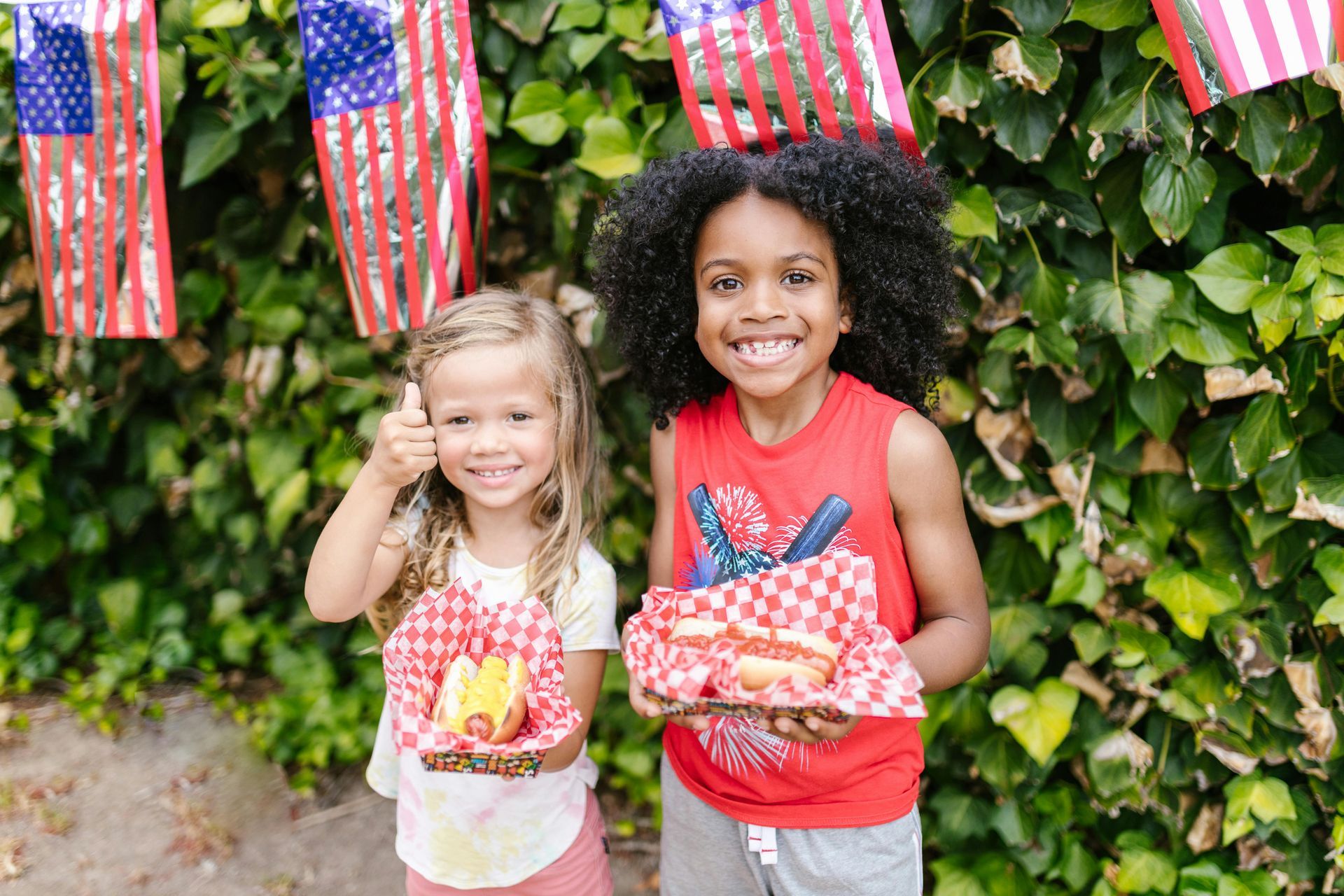 Two kids smile, holding hot dogs, with flags in the background. One gives a thumbs up.
