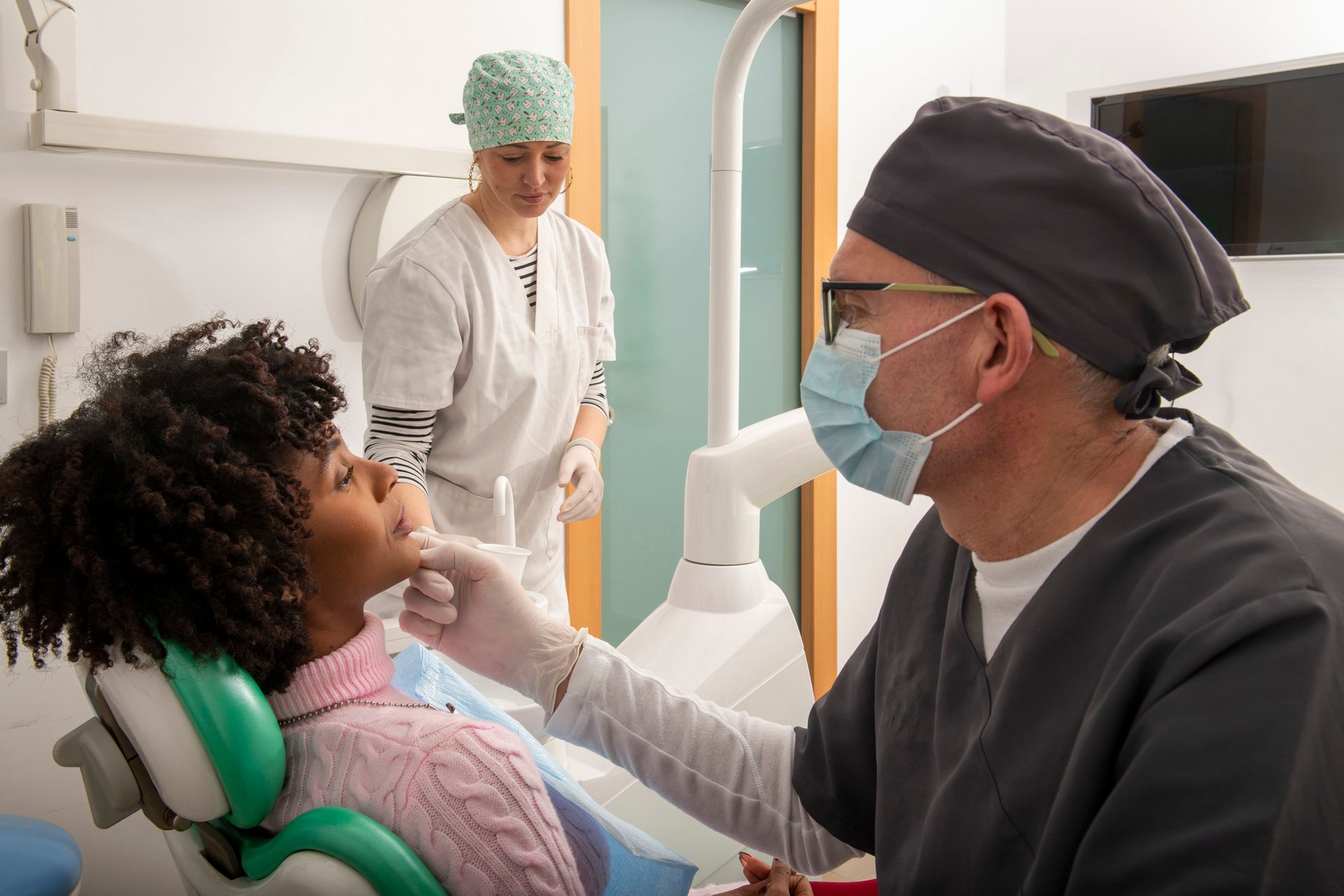 General dentistry exam with masked dentist inspecting female patient’s teeth while assistant stands nearby.