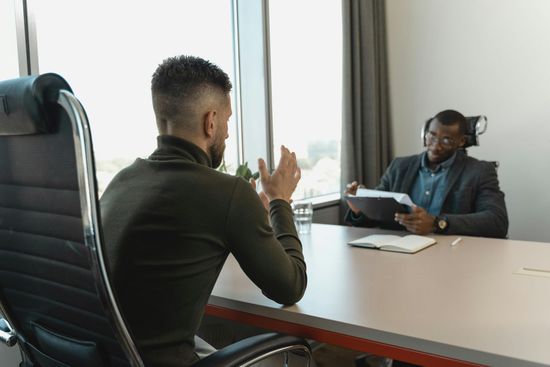 Two people in an office meeting, one seated across a desk holding papers and speaking to the other.