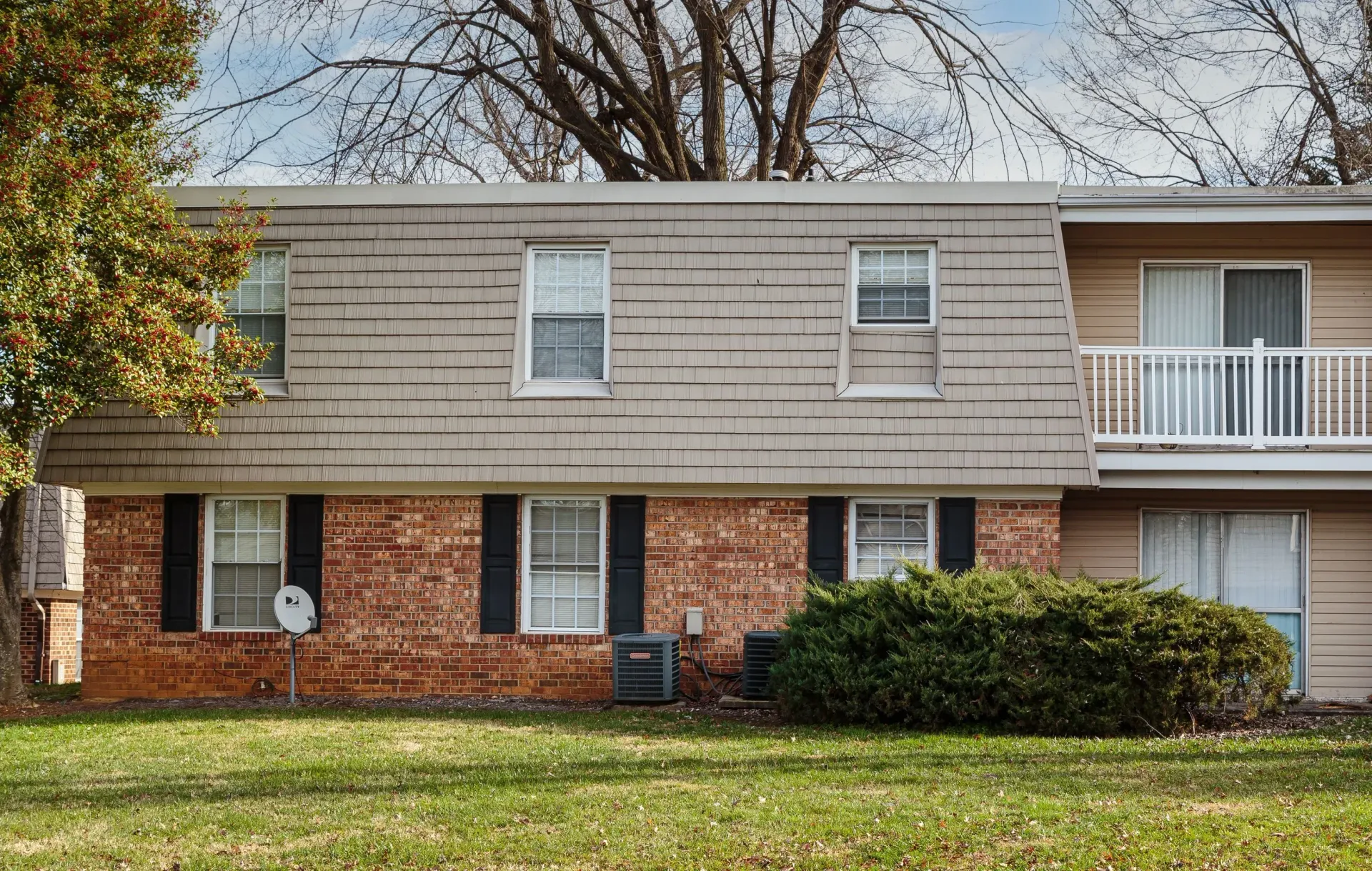 Two-story brick townhouse with tan siding, white shutters, and a small balcony, viewed from the front yard