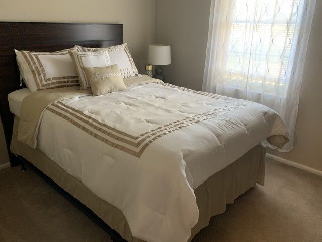 Bedroom with a made bed, white and beige bedding, and a window with sheer curtains.