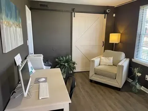 Modern home office with desk, armchair, floor lamp, and white barn door against dark walls.