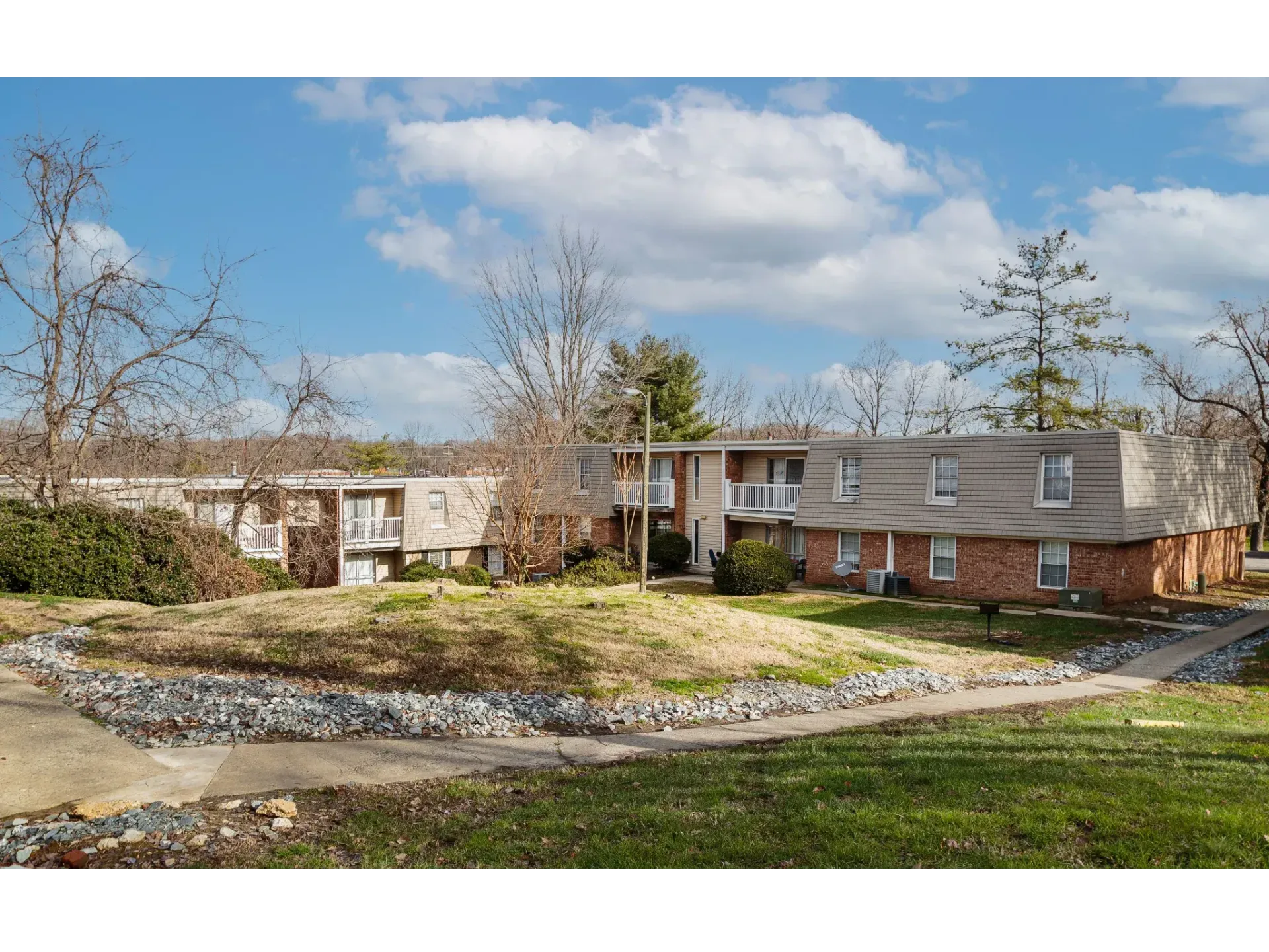 Suburban apartment buildings with brown siding, leafless trees, and a grassy courtyard under a cloudy sky