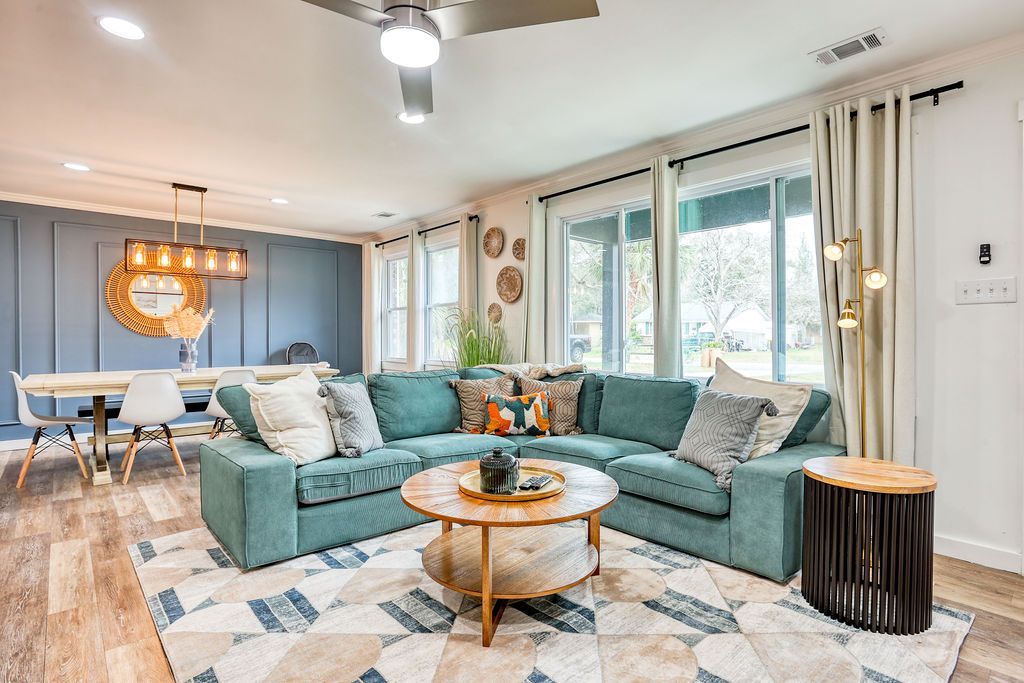Living room with teal sectional, wooden coffee table, dining table, blue wall, and large windows.