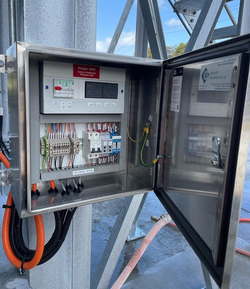 An open, stainless steel electrical control box mounted on a metal structure, containing terminal blocks and breakers.