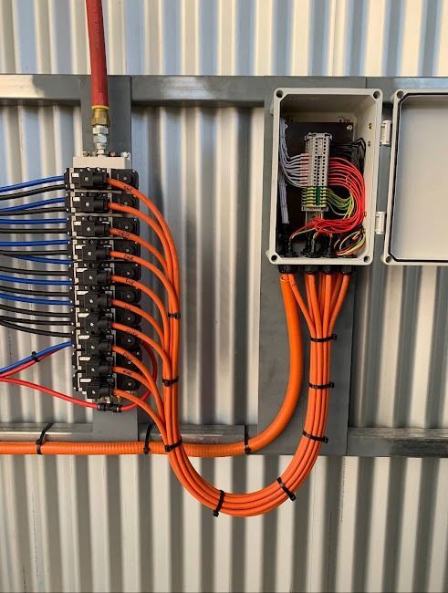 A pneumatic manifold connected to a junction box with organized orange tubing against a corrugated metal wall.