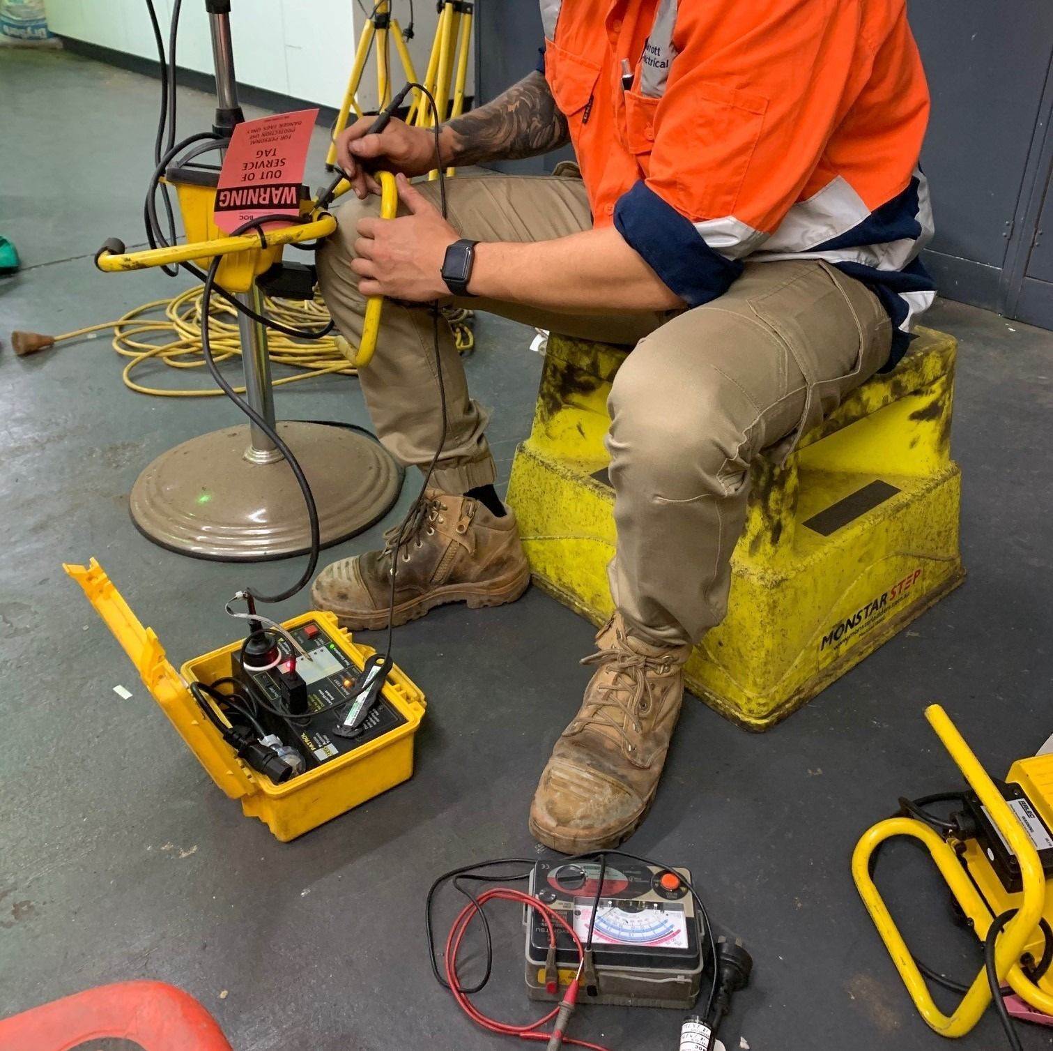 A worker in an orange high-visibility shirt sits on a yellow crate, testing electrical equipment on a workshop floor.