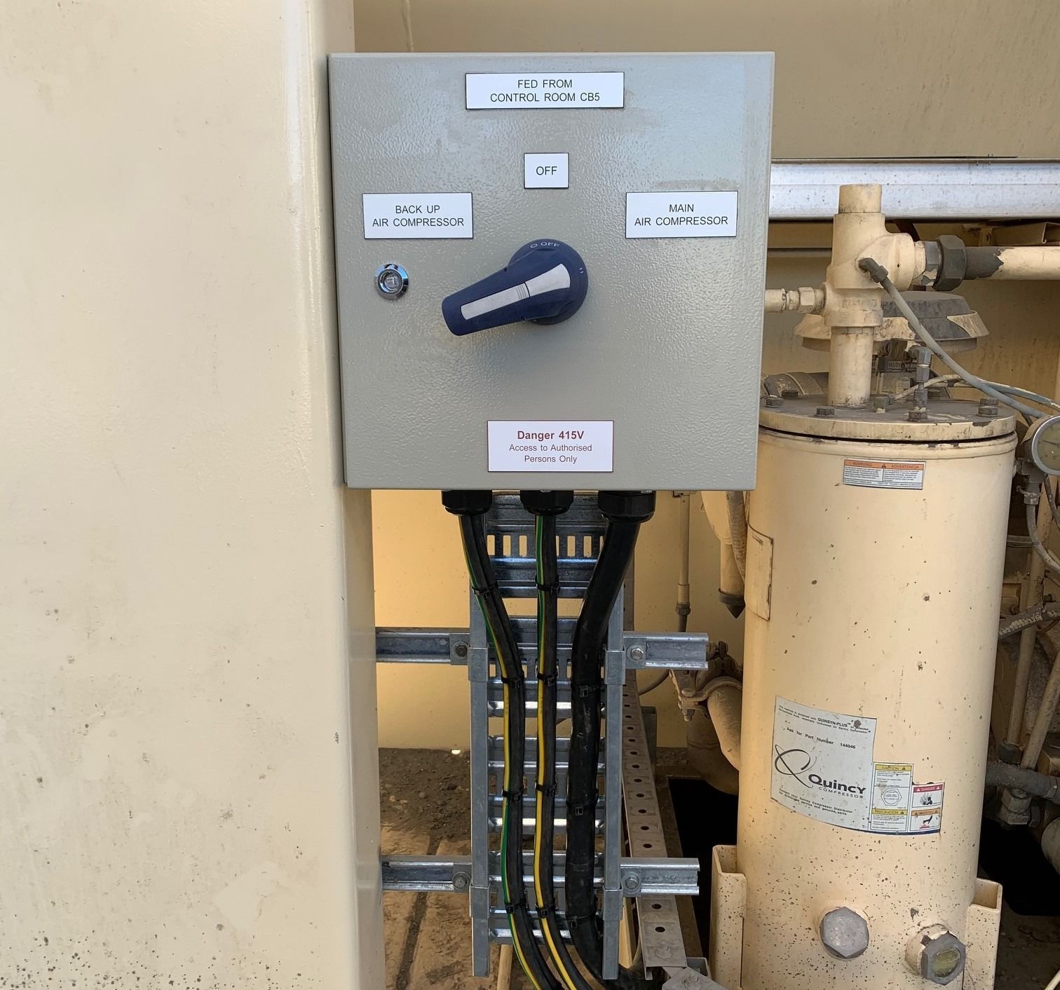 A grey electrical control box with a blue selector switch, mounted next to a beige industrial tank on a metal rack.