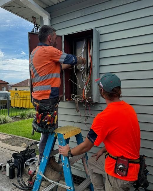 Two workers in high-visibility orange shirts repair electrical wiring in an outdoor wall-mounted box from a ladder.