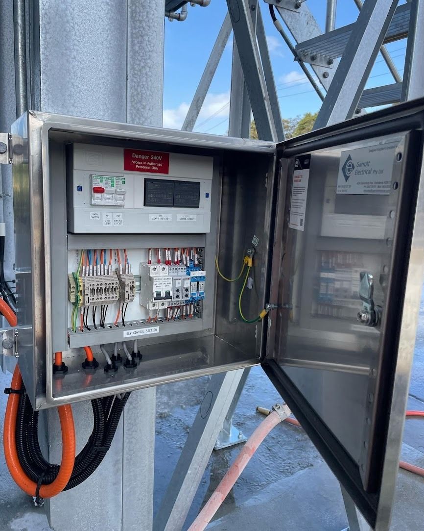 An open stainless steel electrical control box mounted on a metal structure outdoors, showing internal wiring and meters.
