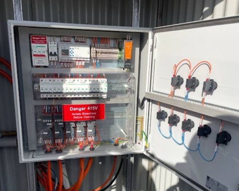 An open electrical distribution box with circuit breakers, internal wiring, and status lights mounted on a metal wall.