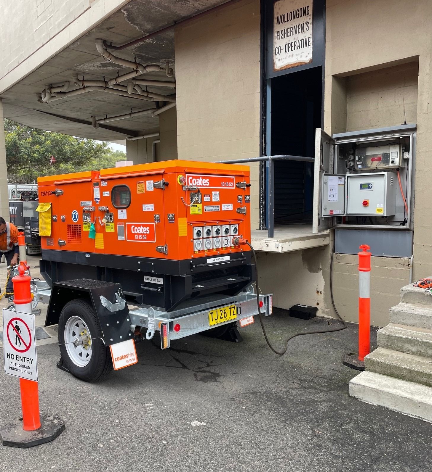 An orange Coates generator on a trailer is connected to an open electrical cabinet outside a building near orange bollards.