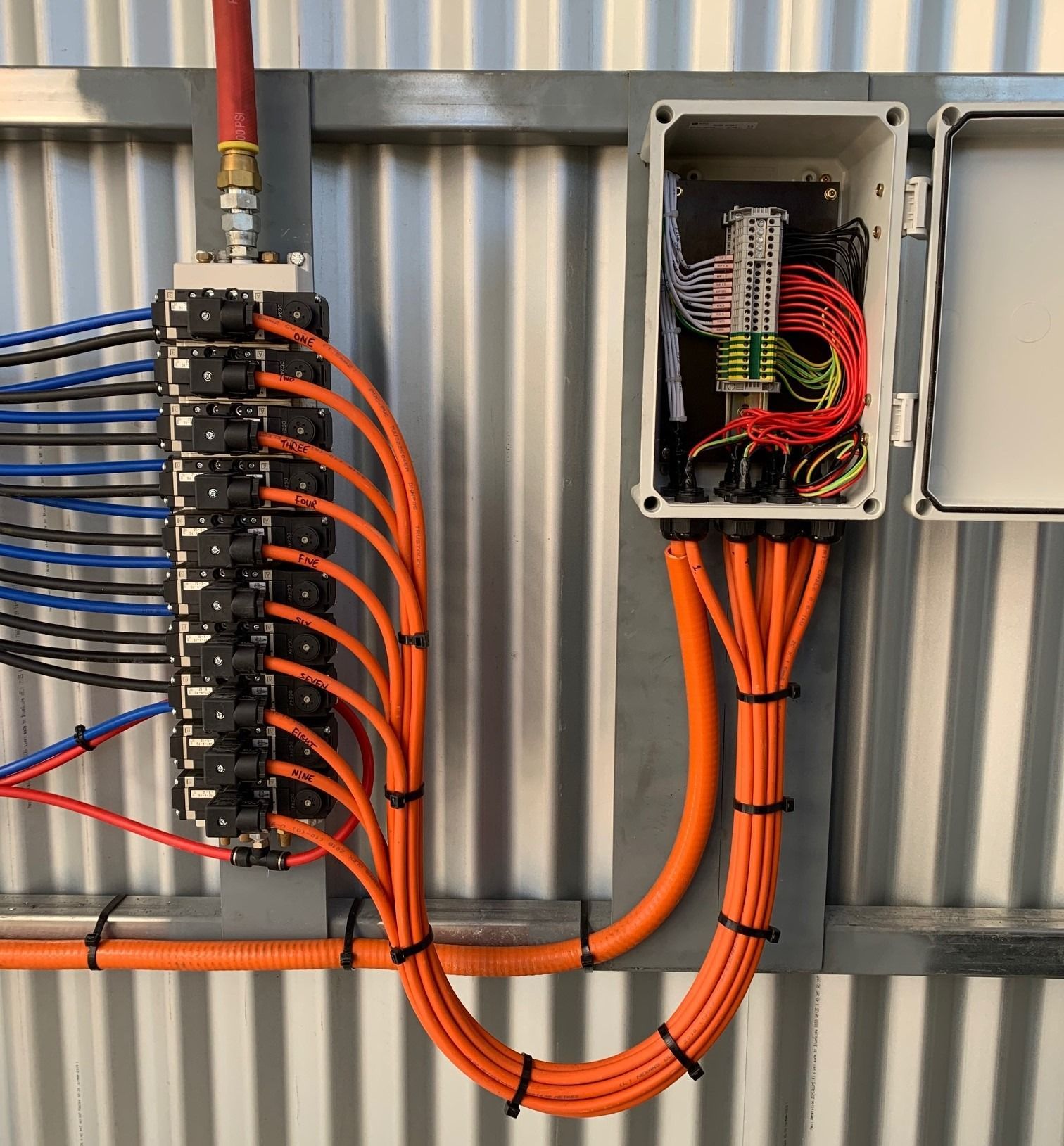 An industrial control box with orange cables connected to a pneumatic manifold mounted on a corrugated metal wall.