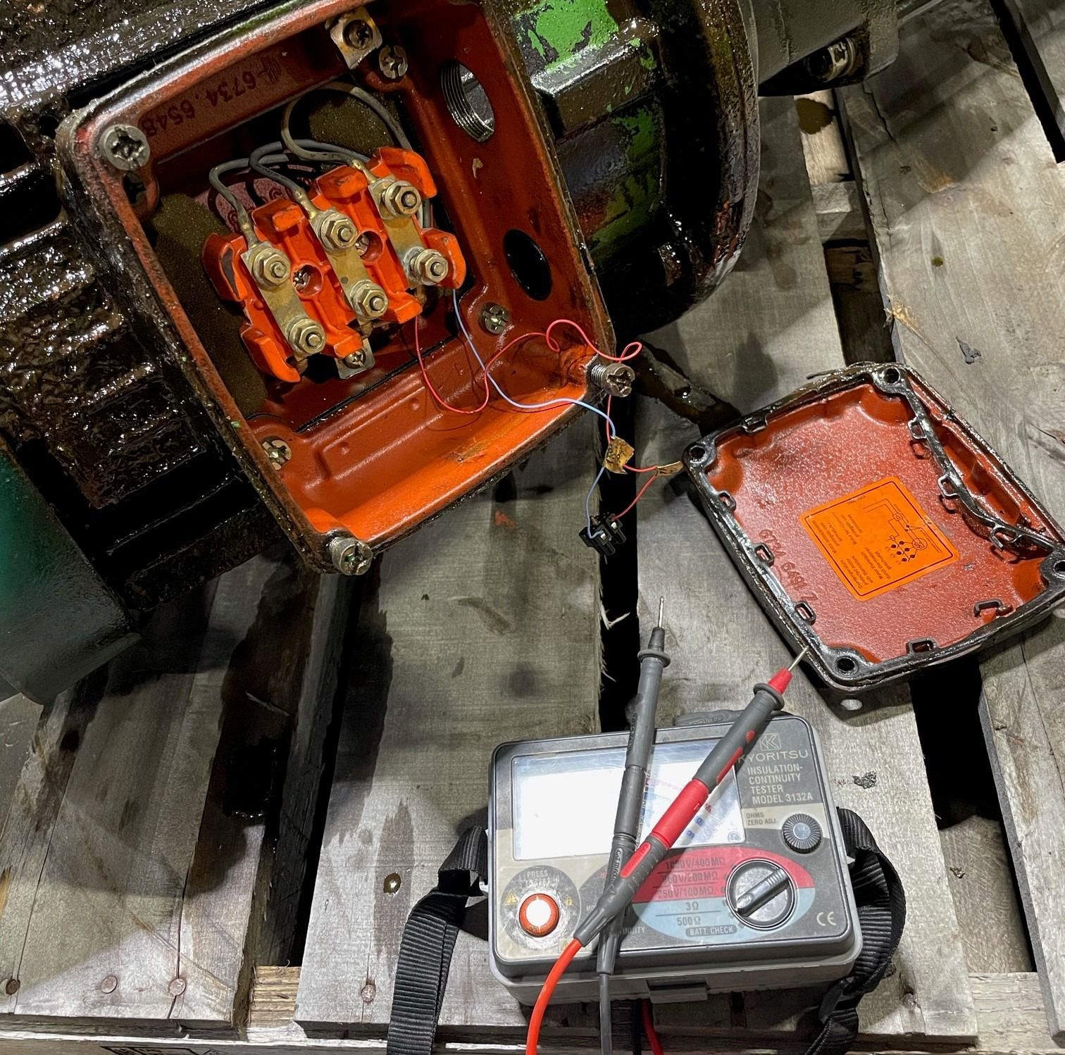 A megohmmeter with red and black leads tests the open terminal box of an industrial electric motor on a wooden pallet.