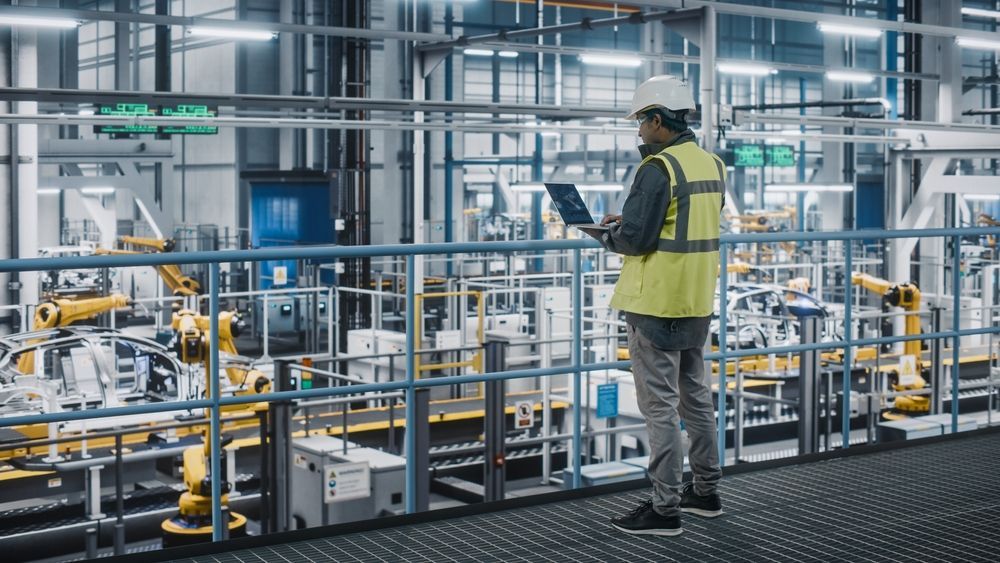 A worker in a high-visibility vest and hard hat stands on an elevated platform, using a laptop in a factory with robots.