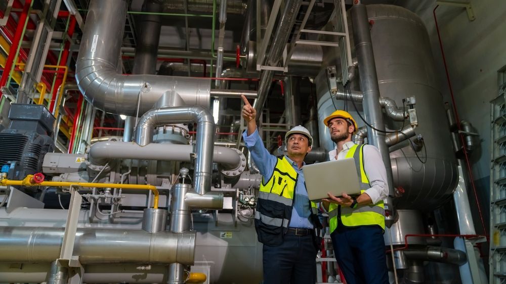 Two people in hard hats and high-visibility vests consult a laptop while inspecting industrial machinery in a plant.