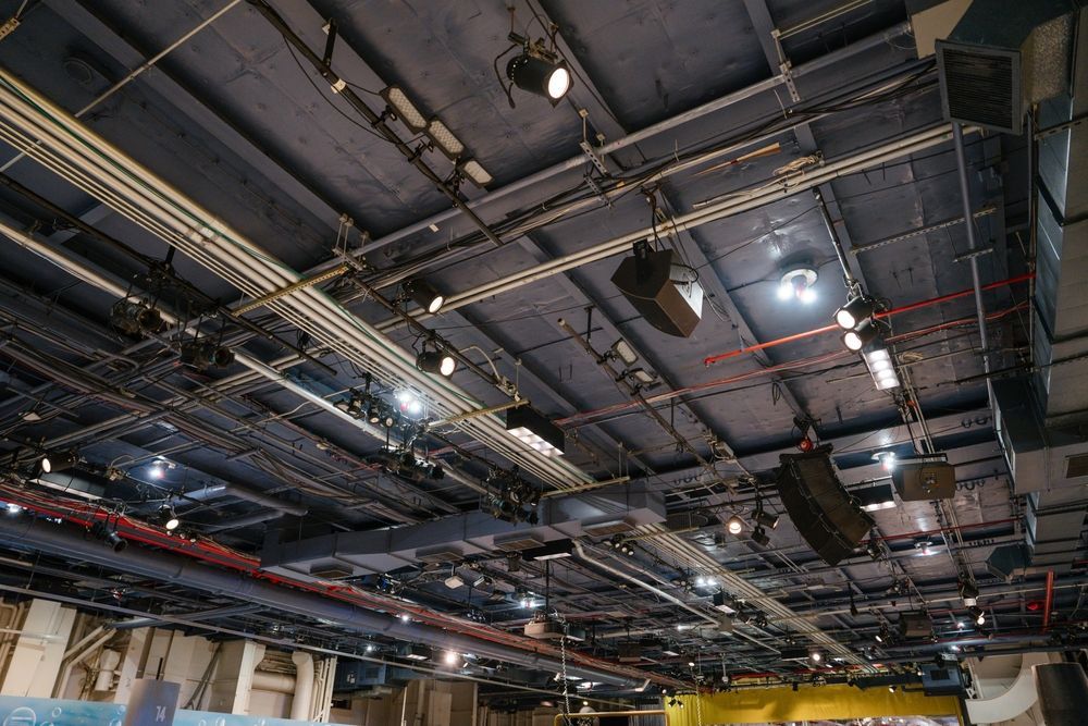 A dark, industrial ceiling featuring complex electrical conduits, ventilation ducts, and a grid of hanging theater lights.