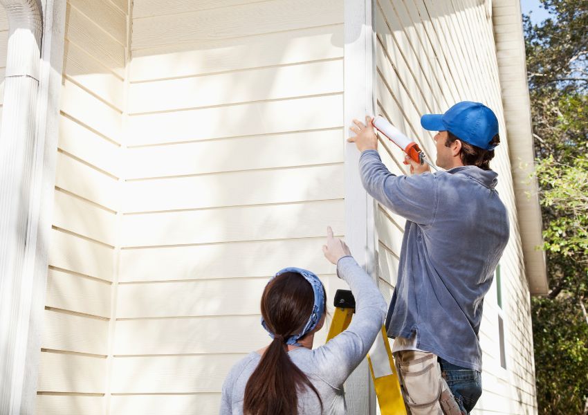 Two property managers face a paneled cream house as they paint the paneling white, in line with HOA regulations.