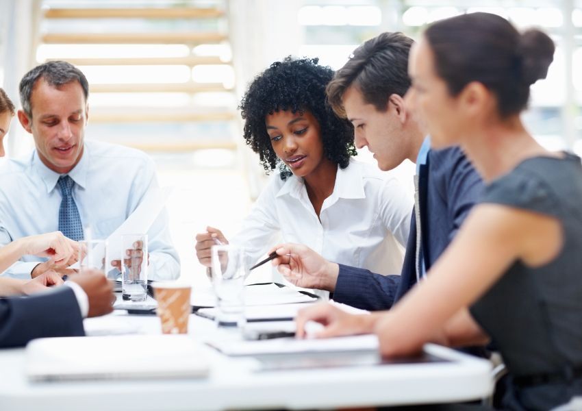 A property manager in a white blouse attends an HOA board meeting with four other members in the frame, discussing policy implementation over a board room table.