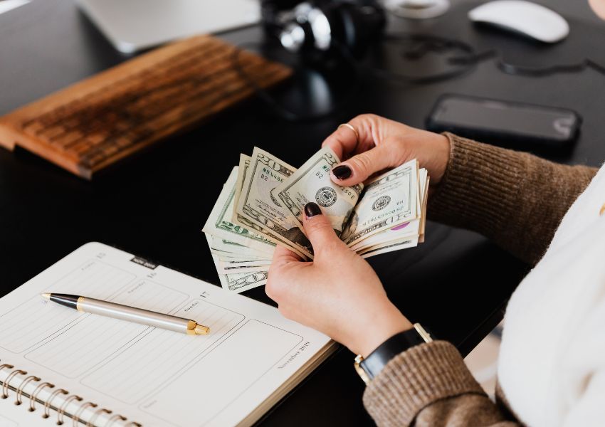A landlord with black nail polish and a watch counts American twenty dollar bills at a desk.