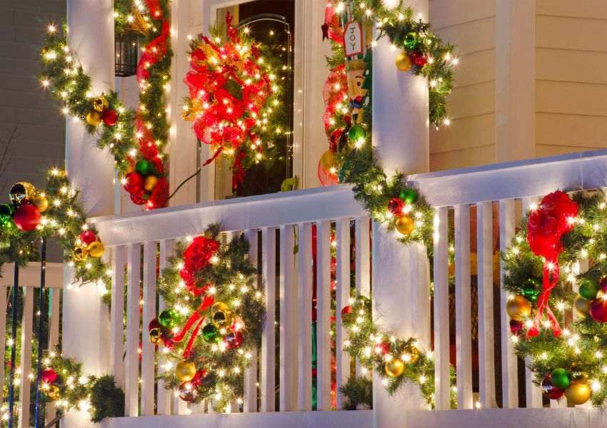 A close up of a pale yellow HOA property with white banisters is decorated with green and red holiday decorations with warm yellow lights and colorful bells throughout.