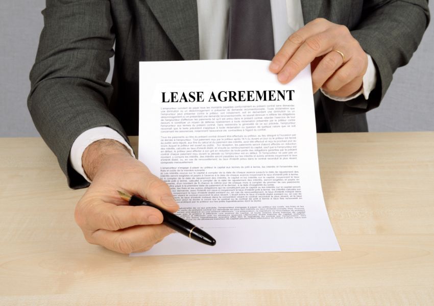 A landlord in a suit holds a lease agreement towards the camera, extending a pen for a tenant to sign.