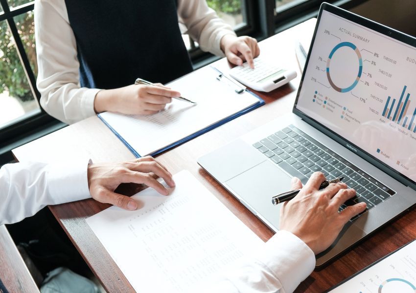 A laptop is pictured on a wooden desk as two property managers create a marketing strategy using the figures depicted.