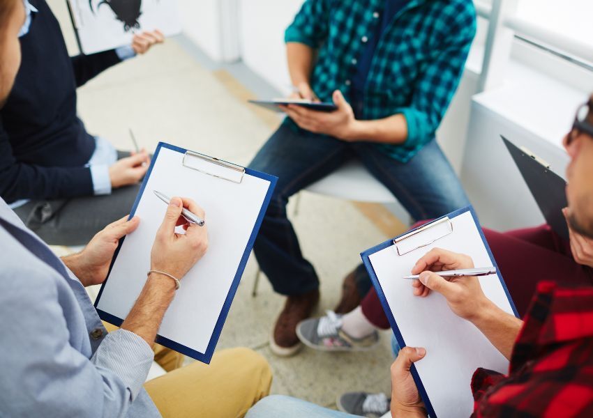 Four property managers hold blue clipboards and silver pens as they sit on chairs facing each other and through HOA checklists and paperwork.