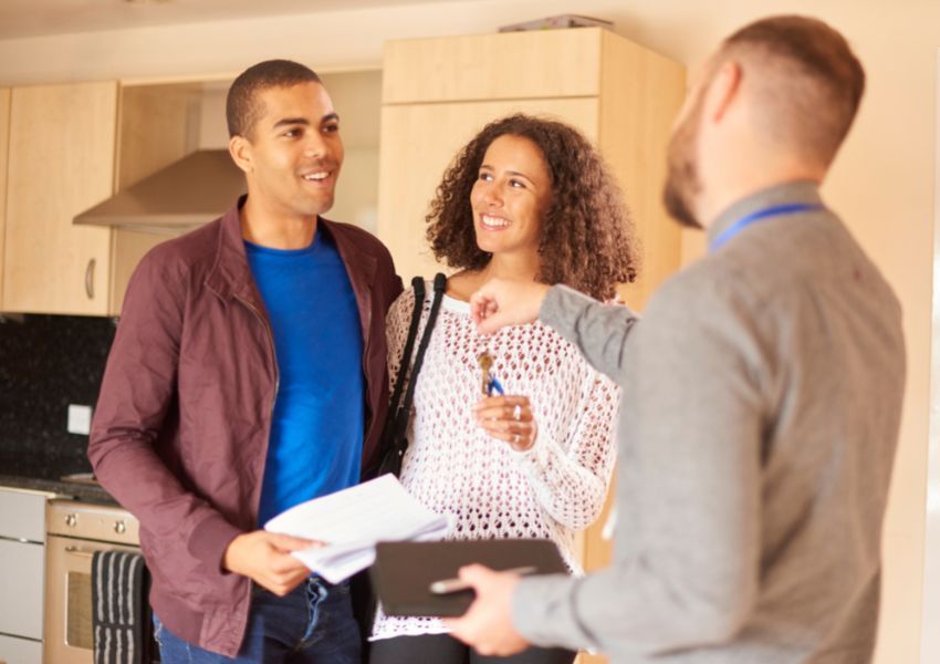 A landlord with a beard and grey dress shirt hands the keys to a rental property to two smiling tenants, inside a kitchen.