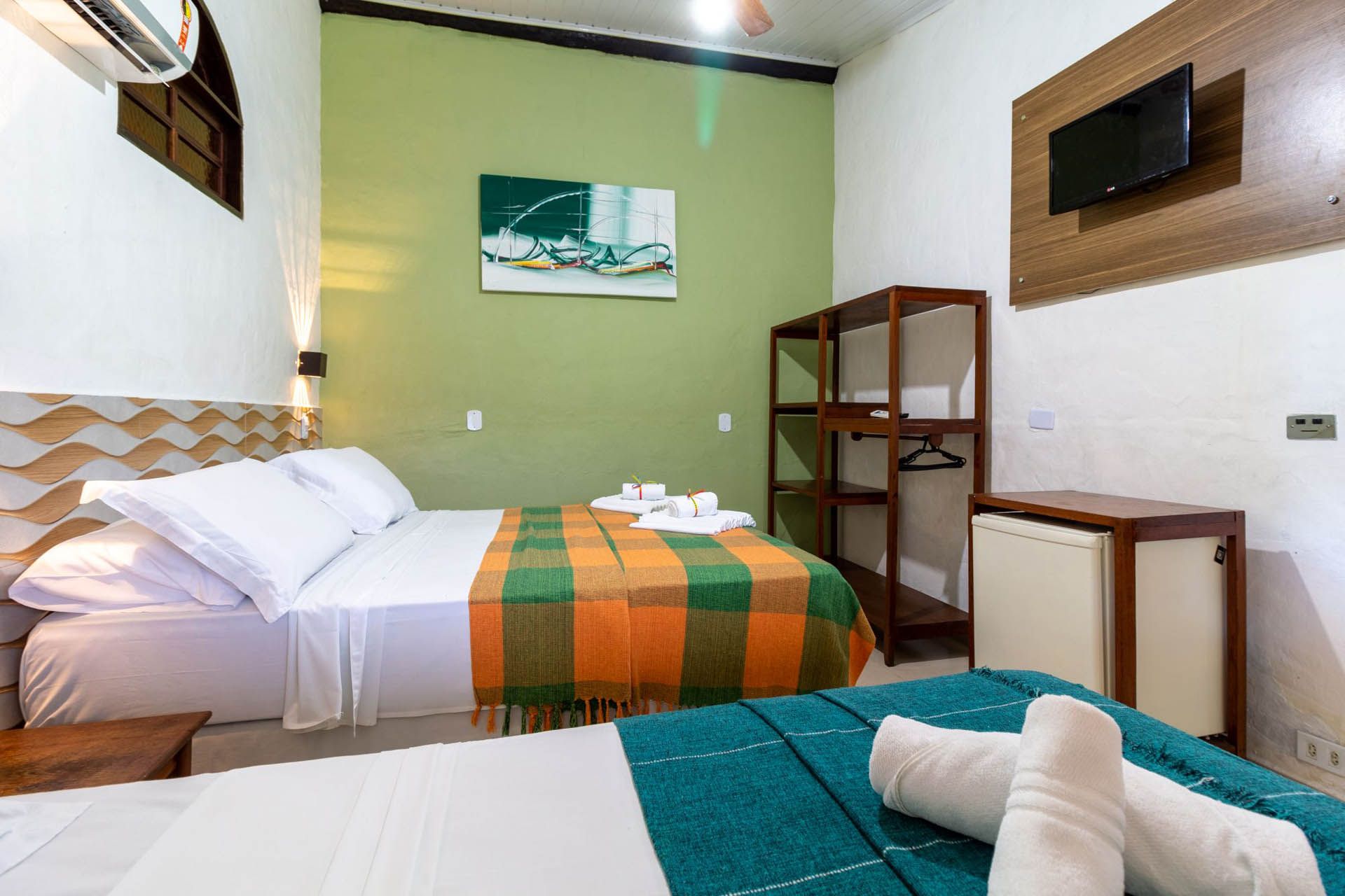 Quarto de hotel pequeno com duas camas, paredes verdes e brancas, prateleiras de madeira e uma janela.