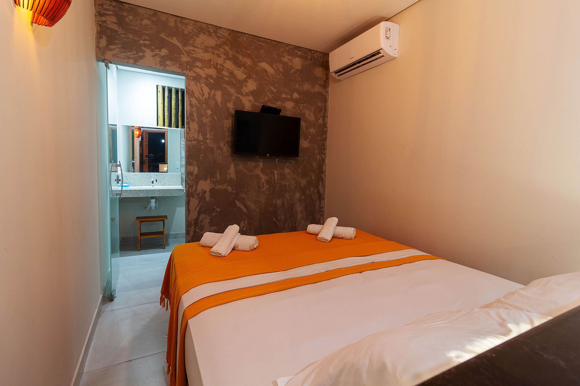 Quarto de hotel pequeno com colcha laranja, TV de parede, ar condicionado e banheiro visível pela porta.