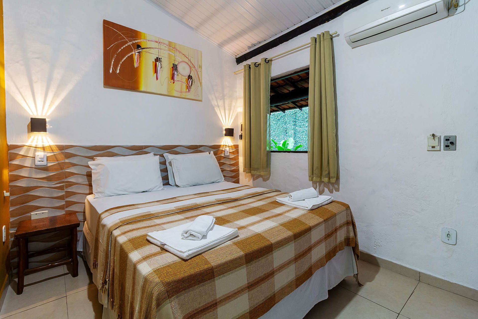 Quarto aconchegante com cama de casal, abajures, obras de arte na parede e janela aberta com vista para a vegetação.