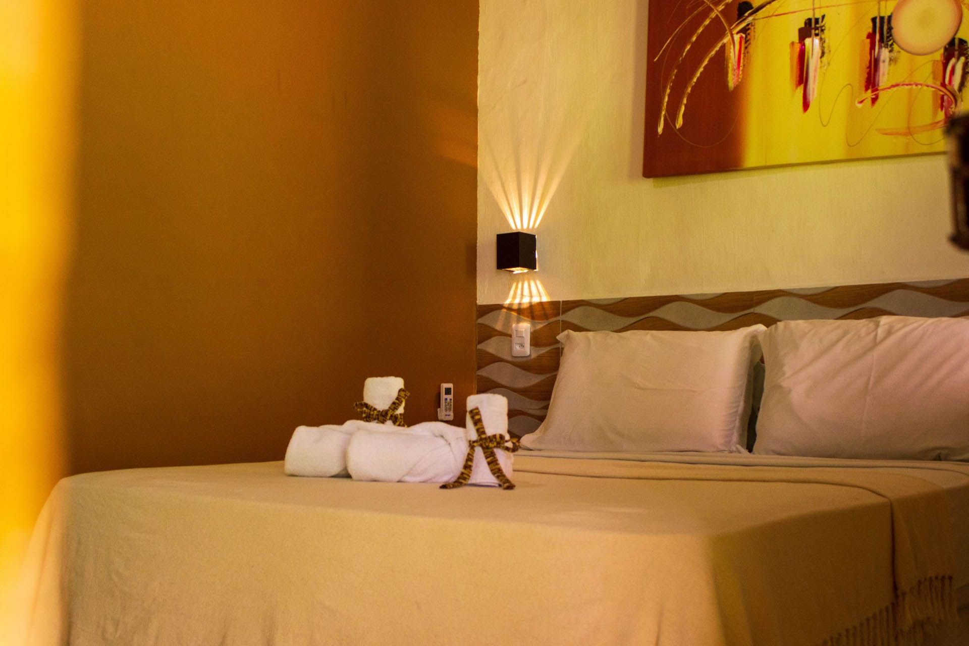 Quarto de hotel aconchegante com cama arrumada, toalhas enroladas, abajur e obras de arte em tons de amarelo na parede.