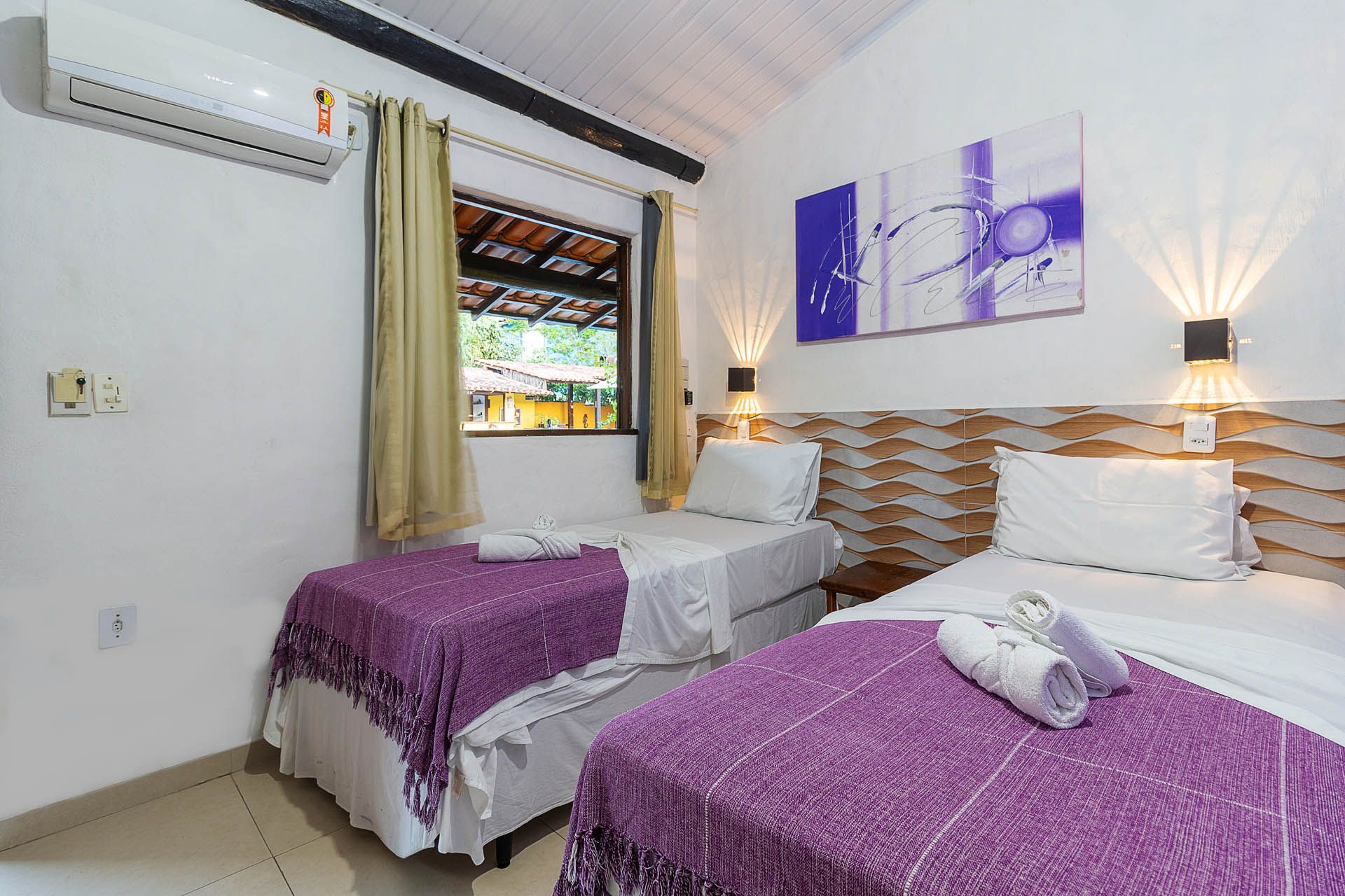 Quarto de hotel luminoso com duas camas de solteiro, colchas roxas, obras de arte na parede e uma janela aberta.