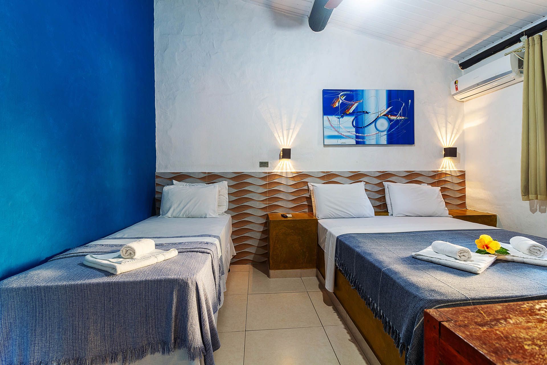 Quarto de hotel com duas camas de solteiro, parede com detalhes em azul, abajures e piso de azulejo.