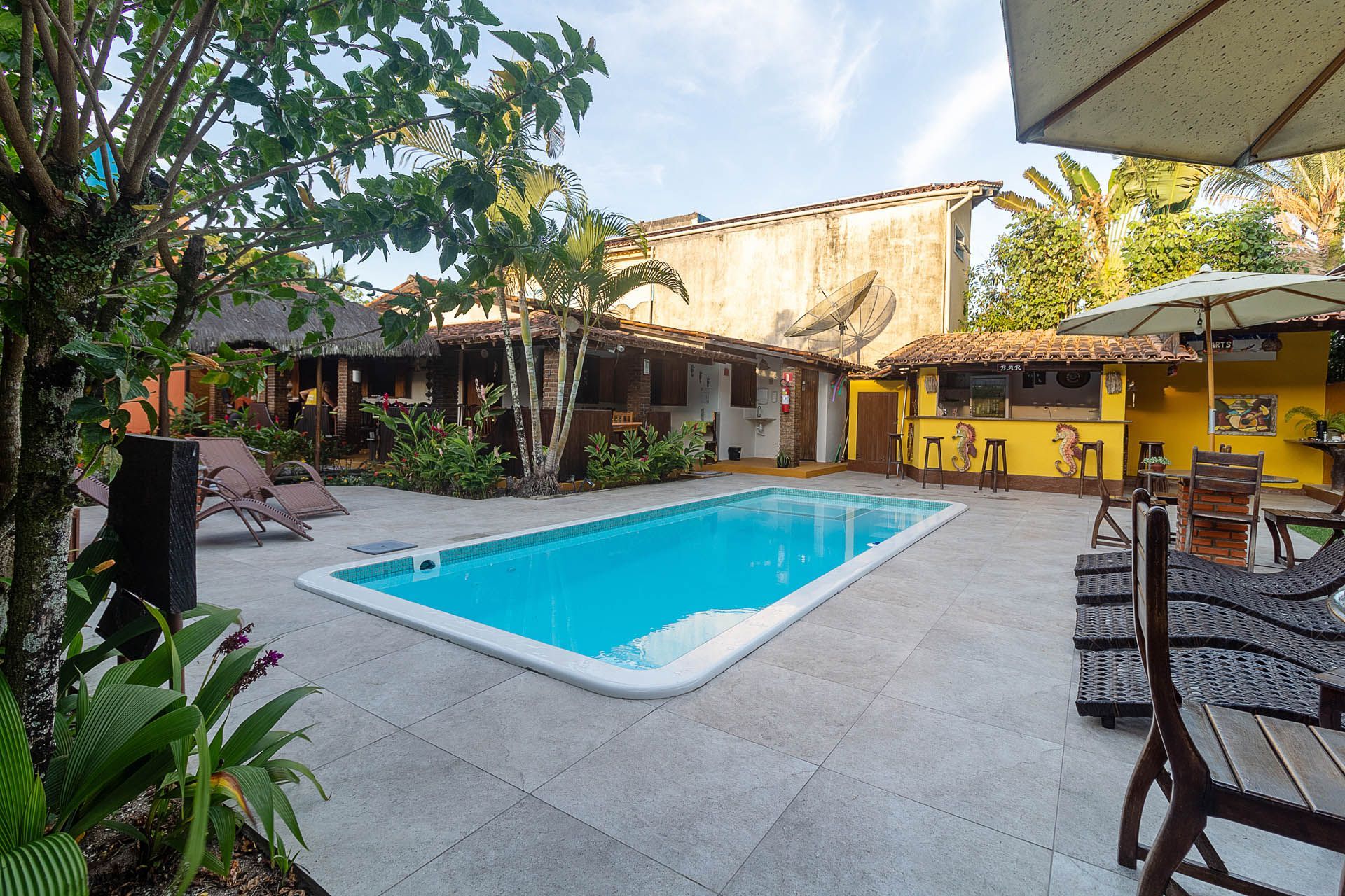 Piscina ao ar livre em um pátio com espreguiçadeiras, árvores e prédios amarelos ao pôr do sol.