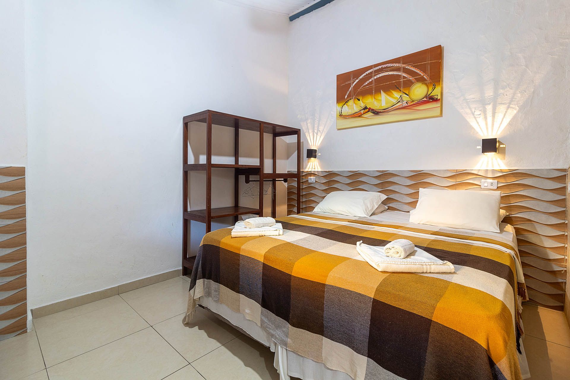 Quarto aconchegante com cama de casal, manta listrada, decoração de parede, luminária e estante aberta.
