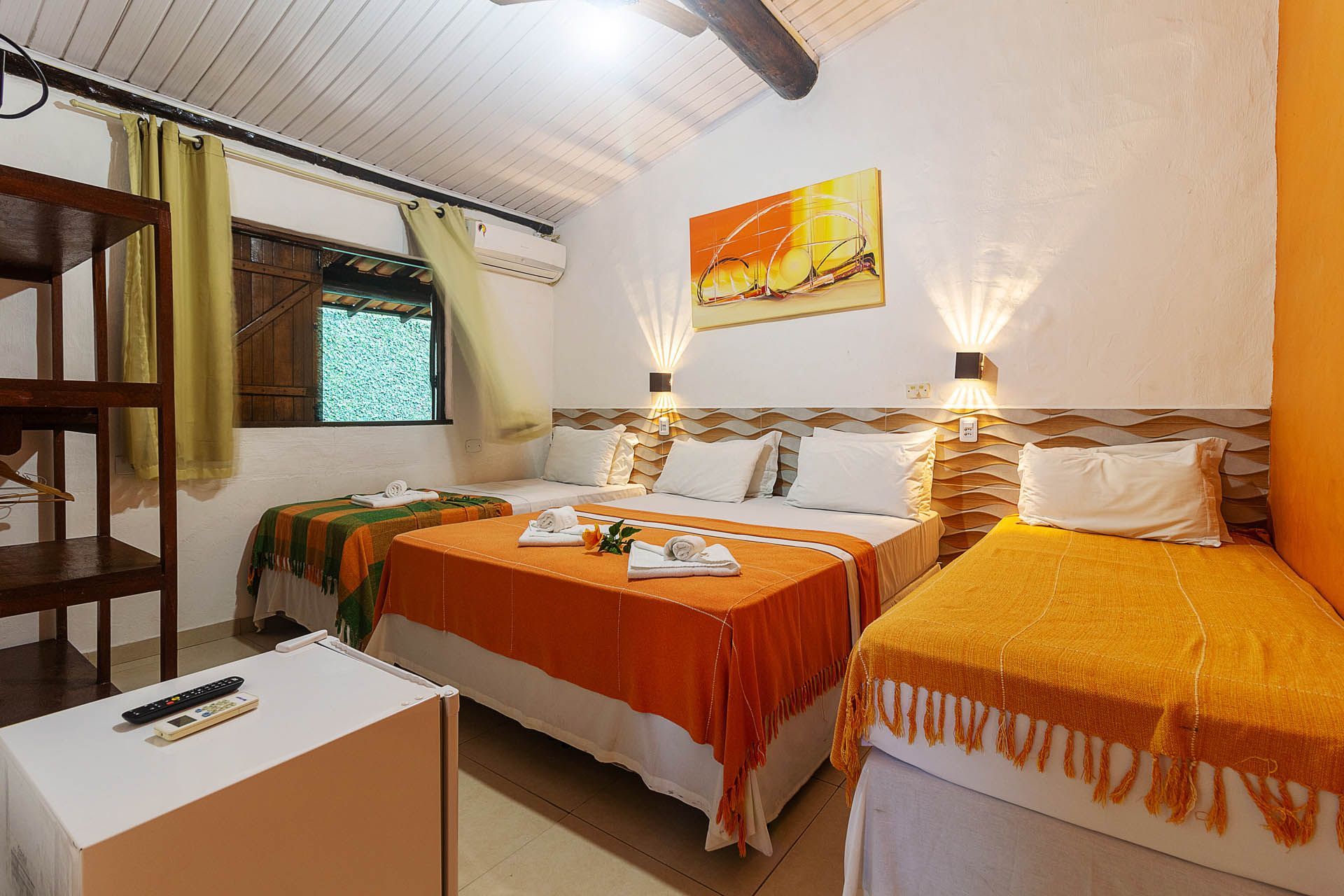 Quarto de hotel pequeno com duas camas cobertas de laranja, paredes brancas, janela e um abajur.