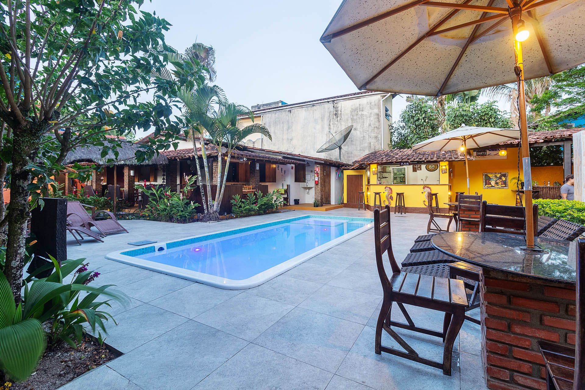 Pátio com piscina azul, terraço de tijolos, cadeiras e guarda-sóis iluminados ao lado de prédios amarelos.