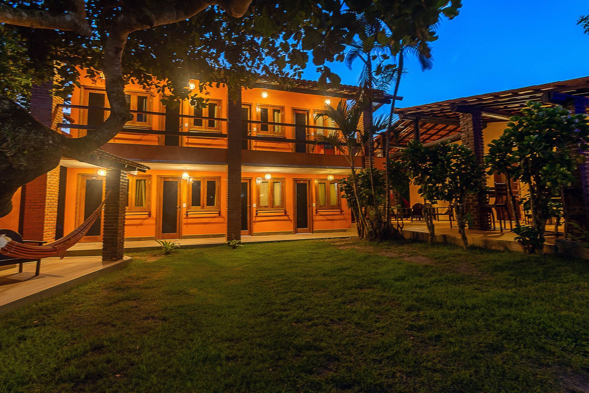 Pátio de um prédio de dois andares com iluminação aconchegante ao entardecer, com árvores, varandas e gramado.