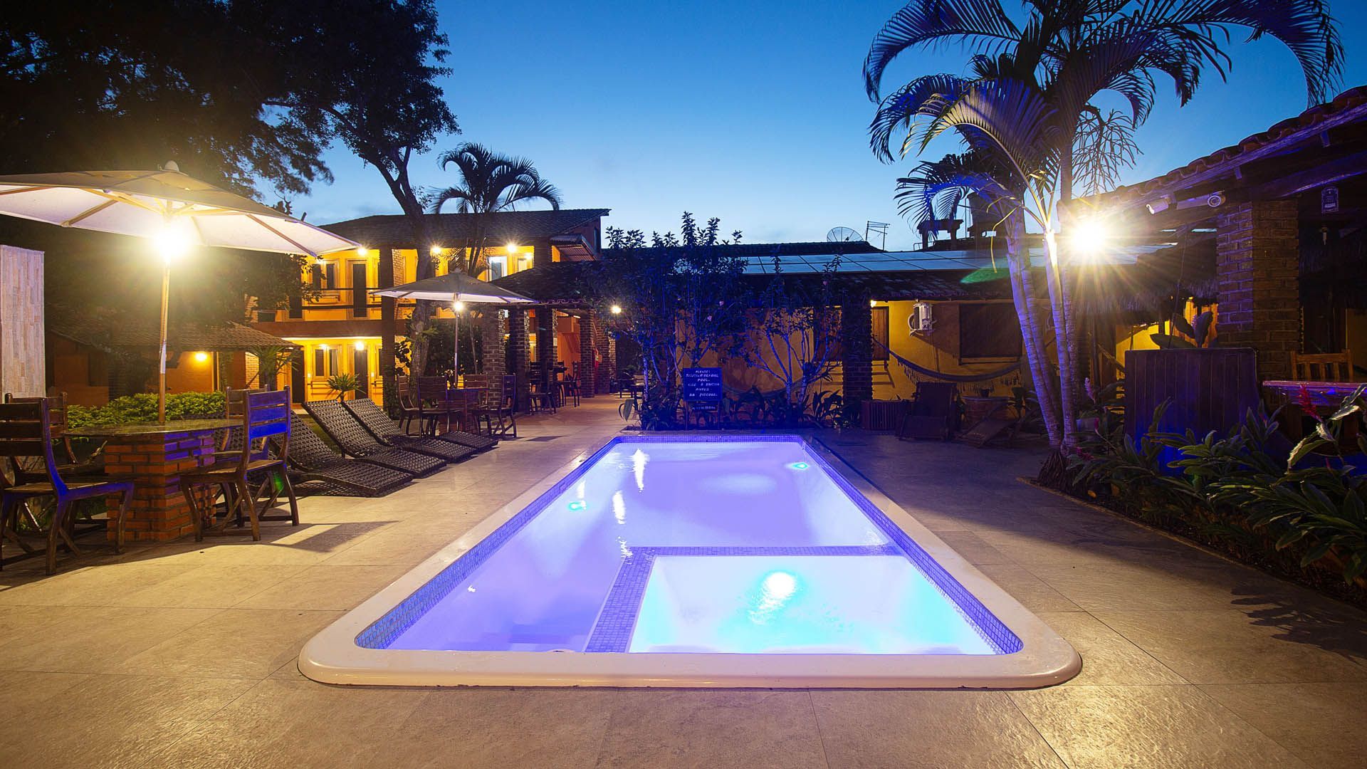 Piscina iluminada no quintal ao entardecer, com água azul brilhante, área de estar no pátio e palmeiras.