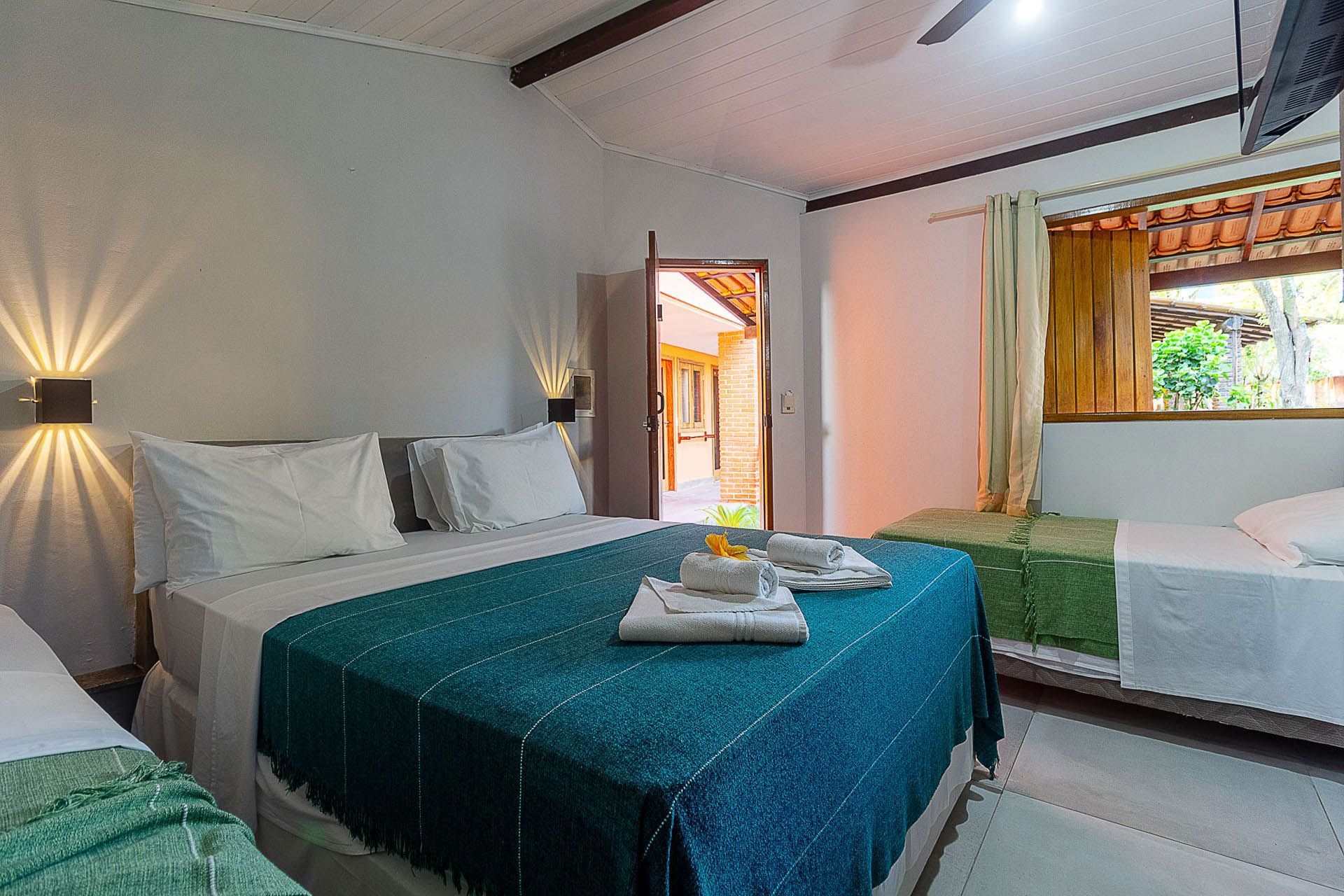 Quarto luminoso com colcha azul-petróleo, roupa de cama branca, apliques de parede e um recanto com assento junto a uma janela ensolarada.