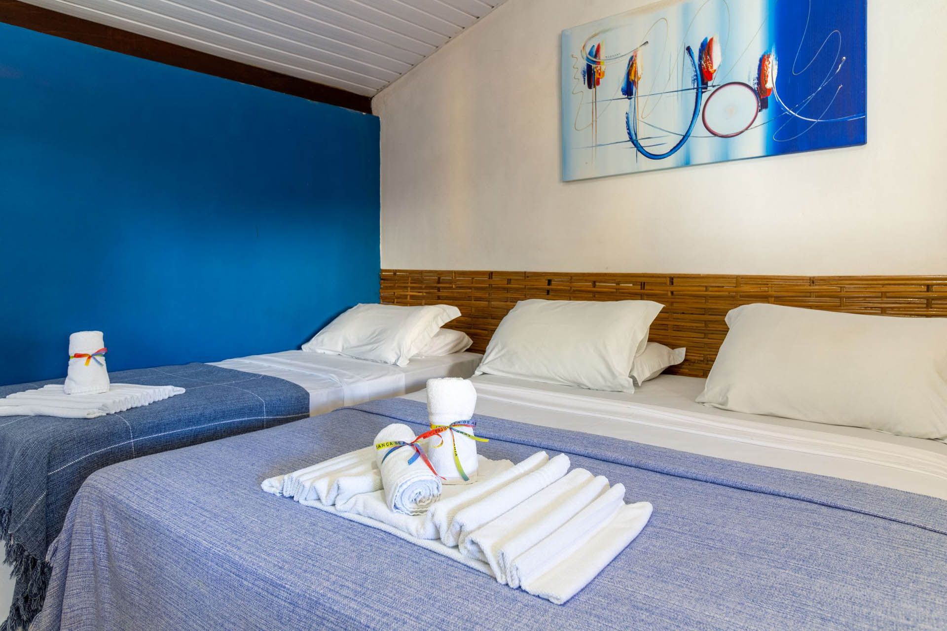 Quarto de hotel com três camas, parede com detalhes em azul e toalhas dobradas na cama do meio.